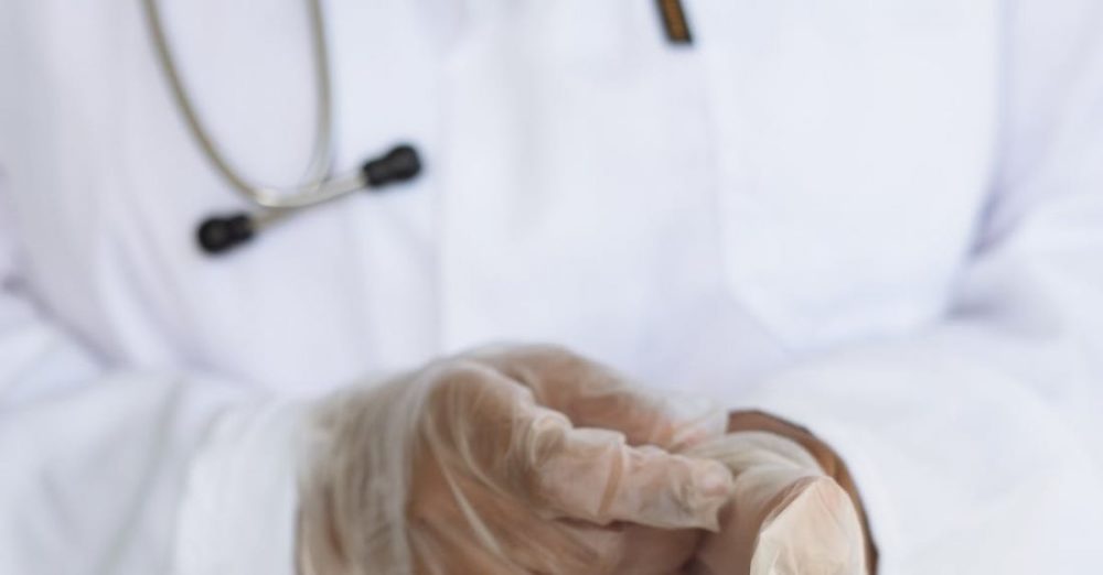 Diagnostics - Faceless ethnic medical worker in lab coat and stethoscope taking of transparent gloves after approaching patients for examination in modern hospital