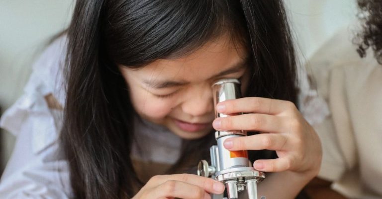 Scientific Research - Cute little Asian girl with long dark hair making experiment with microscope while sitting at wooden table in classroom